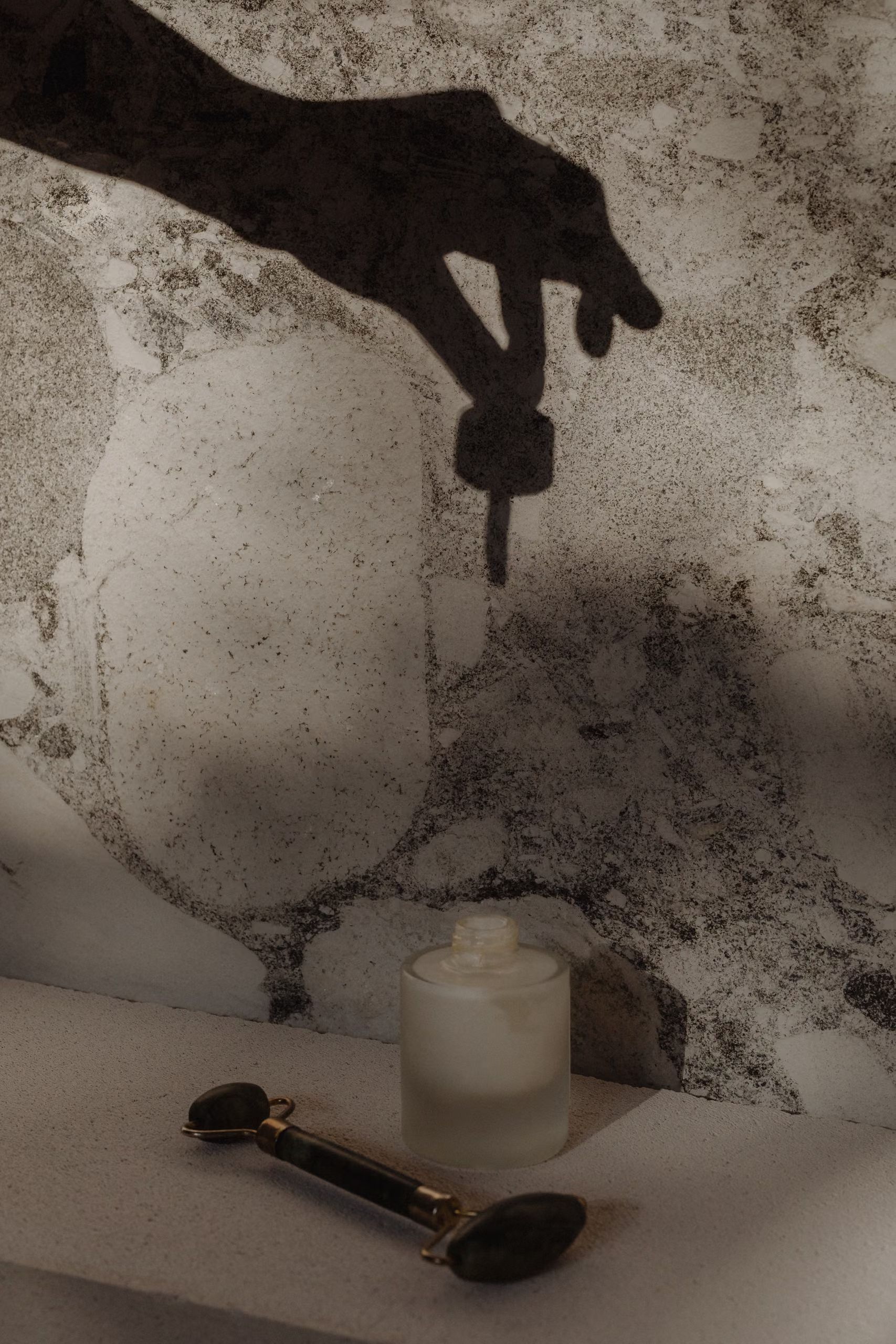 Un flacon de soin en verre et un rouleau de massage en pierre, avec l'ombre d'une main projetée sur le fond.